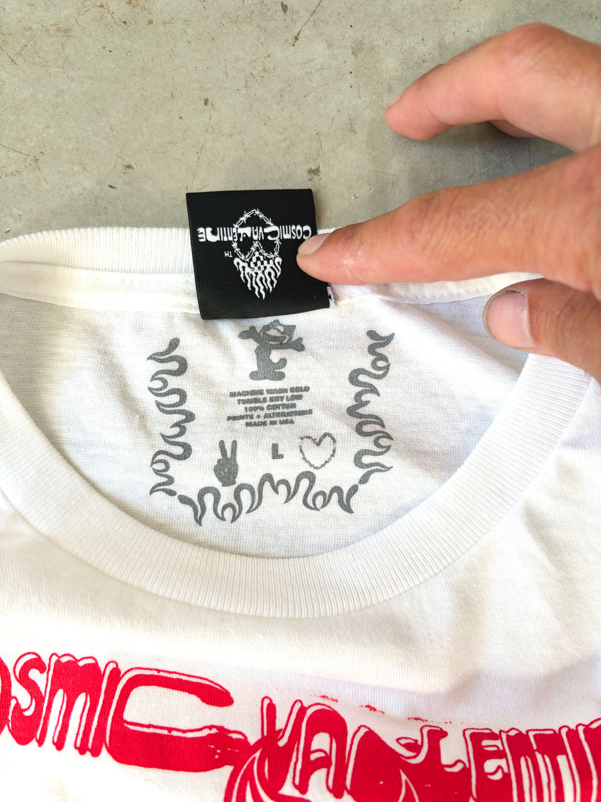 Hand holding the inside collar tag showing the Cosmic Valentine peace sign logo, size L, and printed care instructions