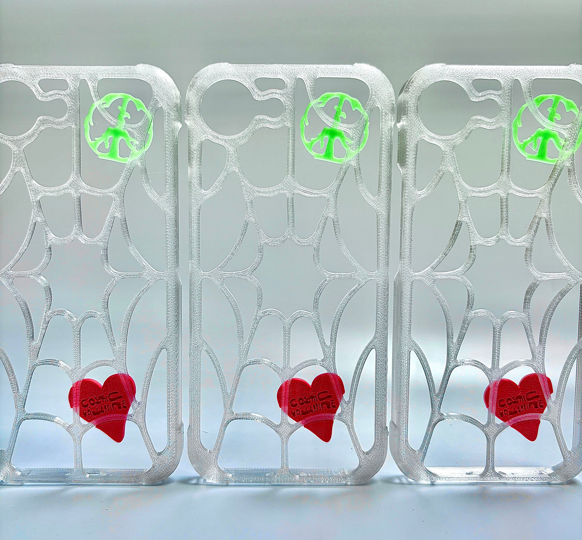 Three clear spiderweb iPhone cases in a row, each with a green peace piece and red heart