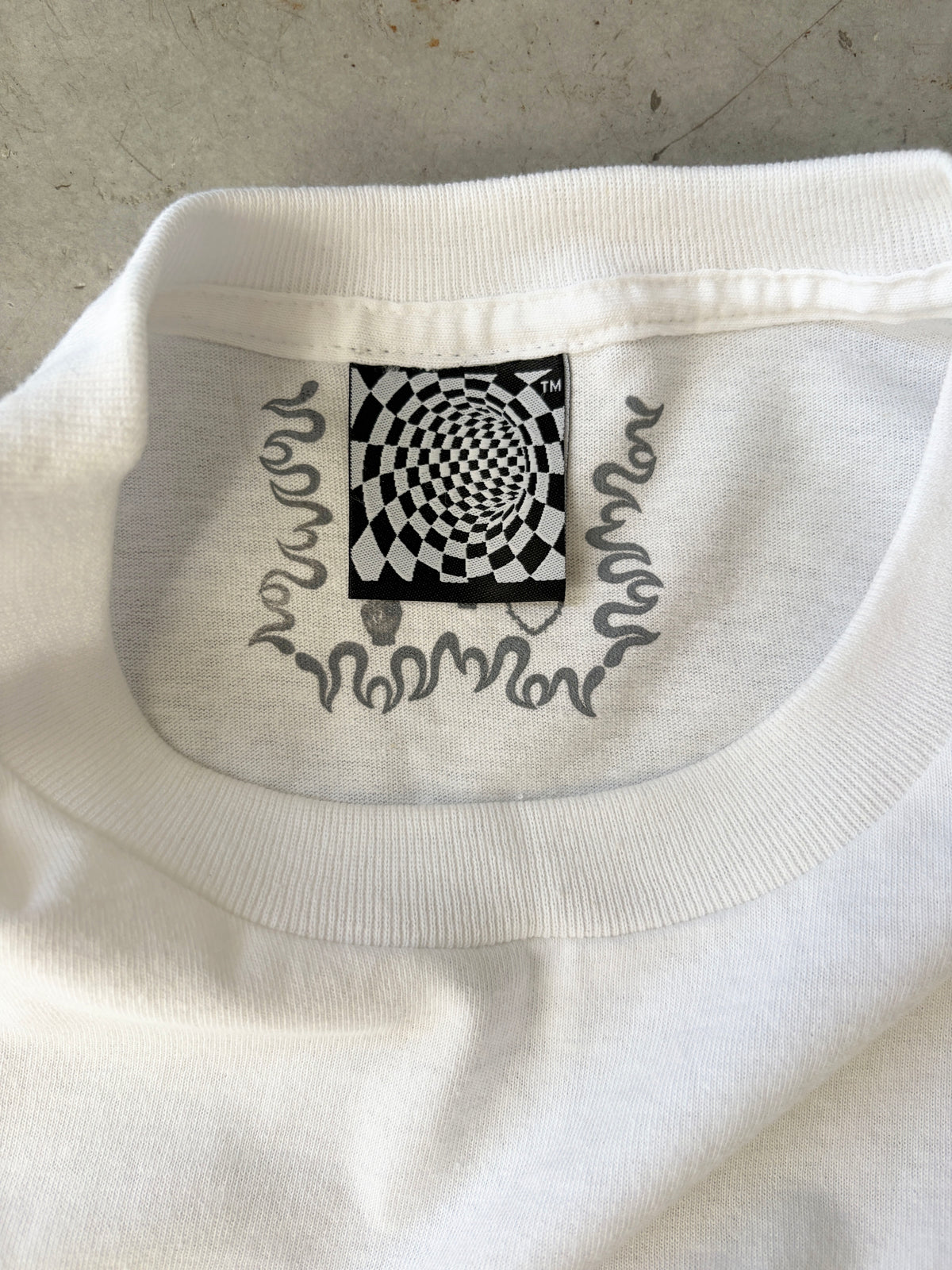 White T-shirt with a black and white checkered spiral design on the neck label and wavy lines printed underneath.