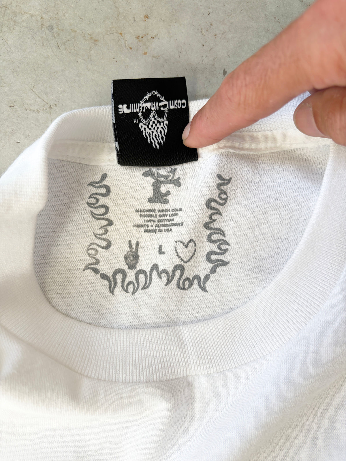 Finger lifting the black woven neck label of the Orbits tee, showing the printed size L tag and Made in USA care text